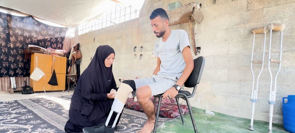 Ibrahim Abdel Nabi, a Palestinian displaced in Gaza, sitting on a chair while his wife helps him wear the handmade prosthetic limb.