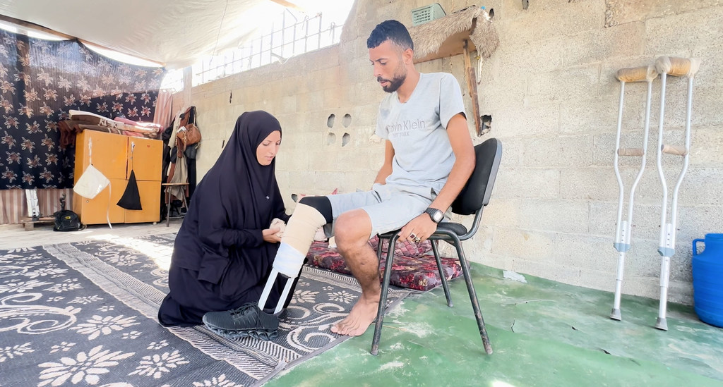 Ibrahim Abdel Nabi, a Palestinian displaced in Gaza, sitting on a chair while his wife helps him wear the handmade prosthetic limb.