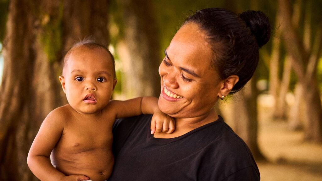 A smiling mother in Palau holds her baby, highlighting community resilience through disaster shelters funded by the United Nations.