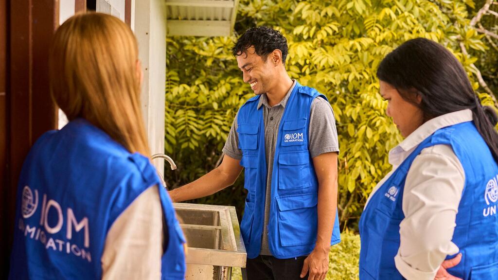 Three IOM staff members in blue vests are shown interacting near a water station, highlighting disaster relief efforts in Palau.
