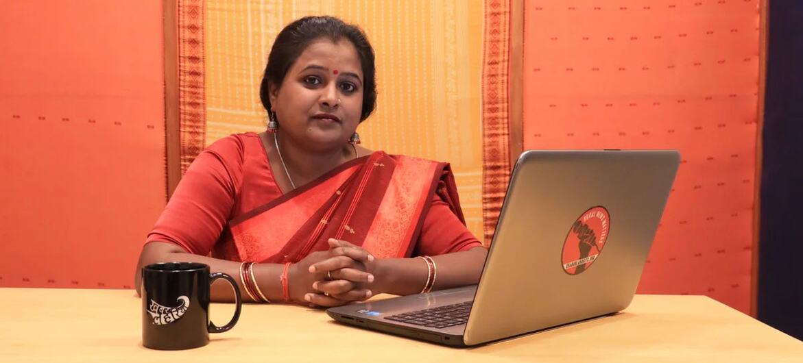 A woman reporter in a red sari sits at a desk with a laptop and a mug, symbolizing grassroots media in India changing narratives on gender issues.