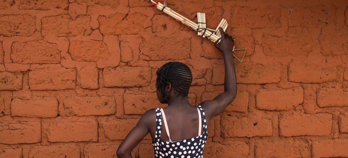 A 13-year-old girl, Elodie, stands with her back to the camera holding a mock weapon, symbolizing her journey from being associated with the anti-balaka armed group to rehabilitation and reintegration in Baoro, Central African Republic.