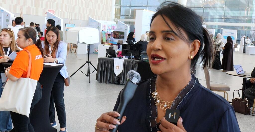 A woman is being interviewed at a business event in Manama, Bahrain, with other attendees and booths visible in the background.