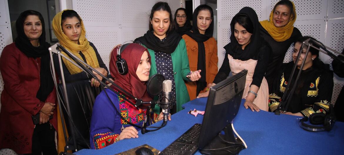 A group of Afghan women in a radio studio. One woman is seated at a desk speaking into a microphone, while others stand around her listening and smiling.