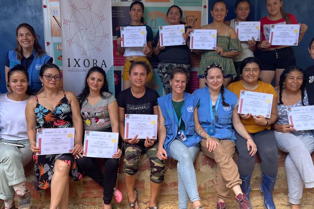 A group of women, including UN Verification Mission in Colombia staff and fashion designers, pose together holding certificates. They are smiling and standing in front of an IXORA Inclusive banner.