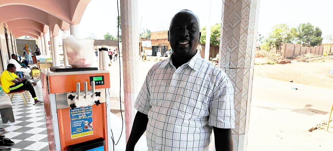 A smiling man, Ibrahim Zakaria Yahya, stands next to an ice cream machine at a shop in Uganda.