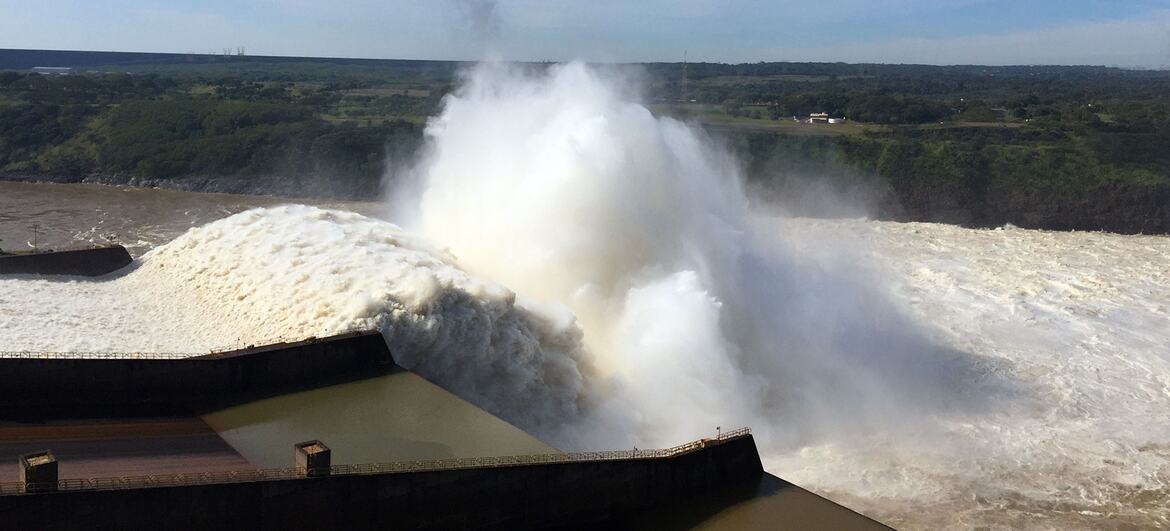 A massive water release from the Itaipu Dam on the Paraná River, creating a huge plume of white water and spray against a blue sky. The dam is jointly operated by Brazil and Paraguay.