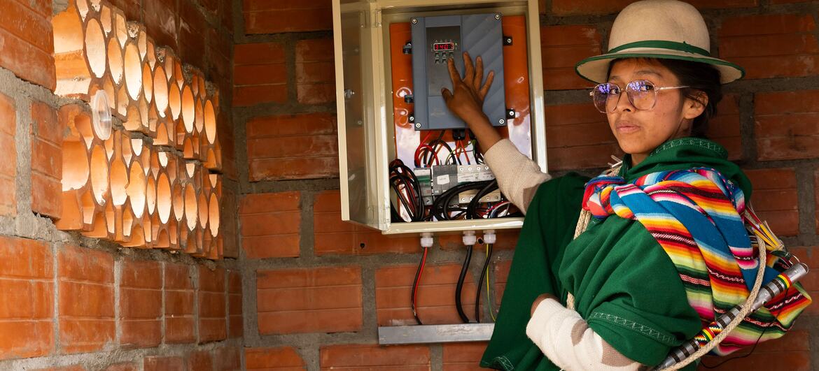 A Bolivian woman in traditional colorful clothing and a hat stands next to an open solar energy control panel mounted on a brick wall, demonstrating the technology's impact on rural communities.